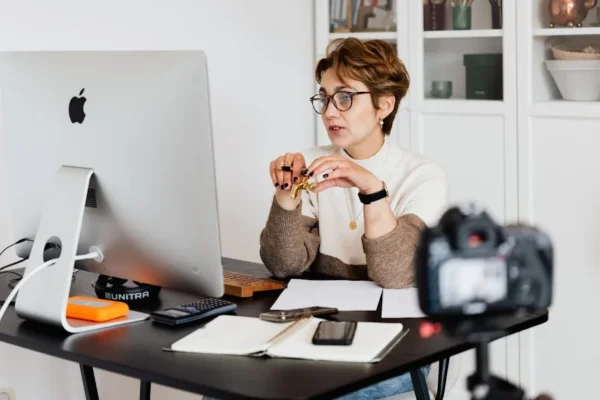 Smart short haired female coach in glasses having web seminar using modern monoblock PC while being recorded on video with camera on tripod in modern study