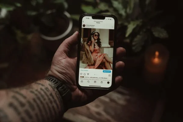 A tattooed hand holding a smartphone displaying an Instagram post indoors.