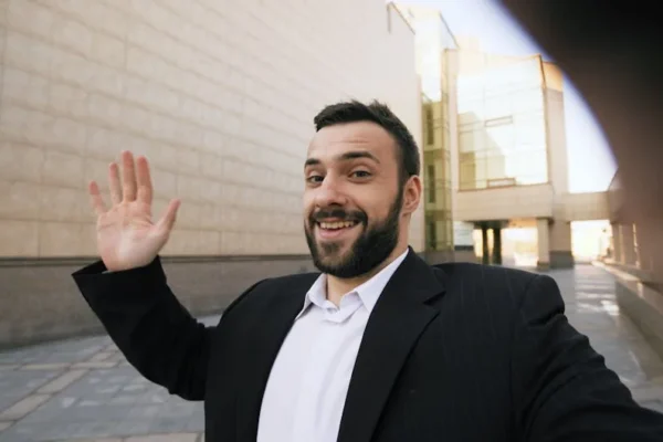 Smiling businessman in suit taking a selfie outdoors by a modern building.