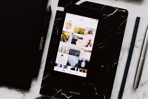 Top view of black notebook with cellphone above with pictures on screen and two pens aside located on marble table