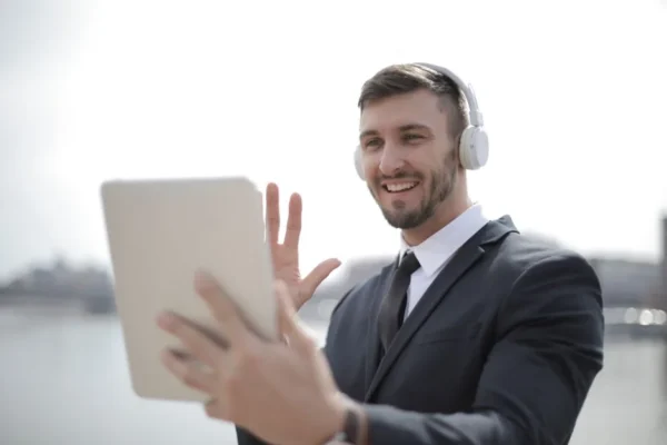 Smiling businessman in suit using a tablet with headphones outdoors, engaging in a video call.