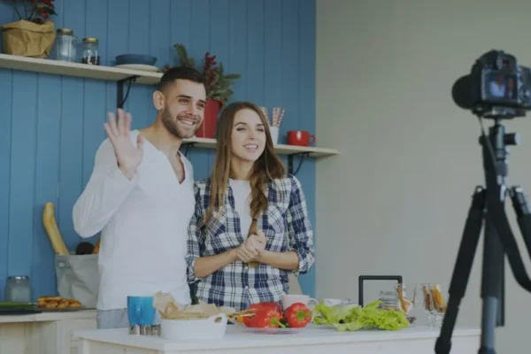 A smiling couple waving and recording a cooking video in their kitchen. Fresh vegetables on the counter.