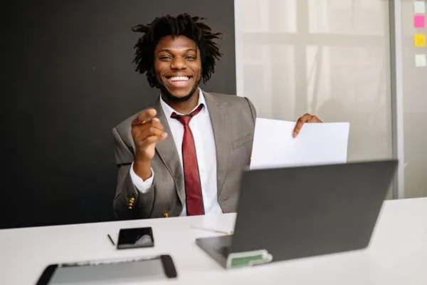 Smiling businessman holding paper at desk with laptop, embodying confidence and professionalism.