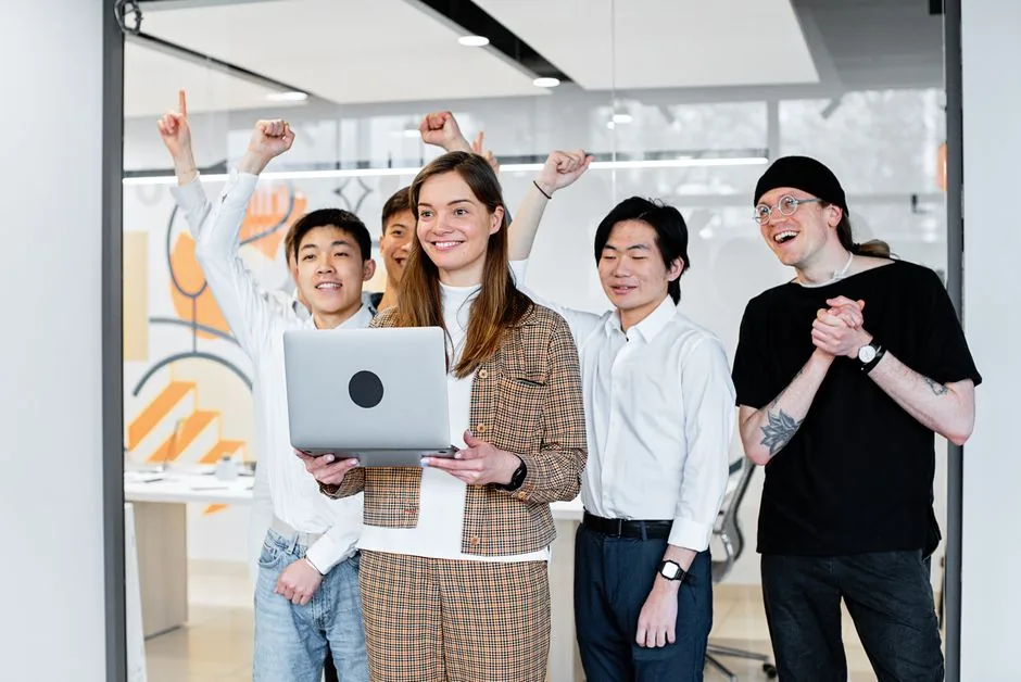 Group of diverse colleagues celebrating success while collaborating on a laptop indoors.